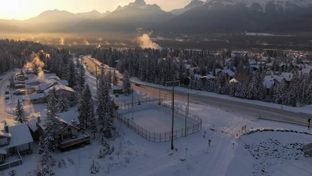Morning sun hits the tips of Rocky Mountains while an outdoor ice skating rink remains untouched. Aerial. Canmore Alberta.
