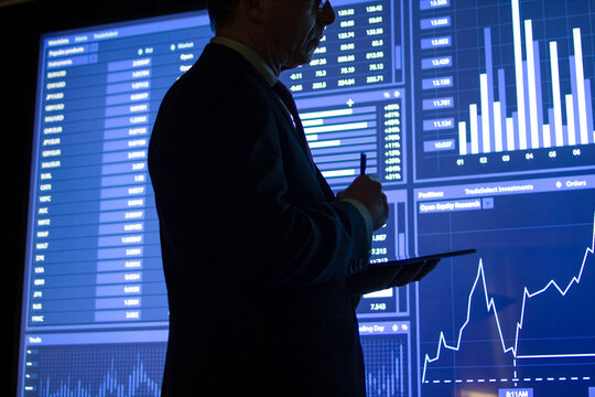 Close-up of employee preparing market data report. Serious man analyzing and calculating numbers from graphs on big digital screen in dark office room. Stock exchange market and finance concept