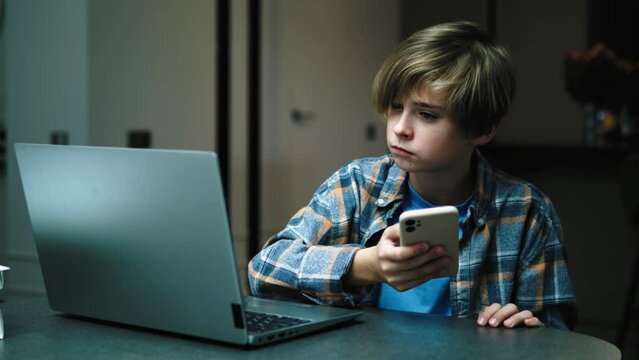 Attractive boy teenager using smartphone mobile phone for chatting, sitting near laptop looking at screen, playing game or doing shopping online. Addicted teenager to gadgets, new generation 