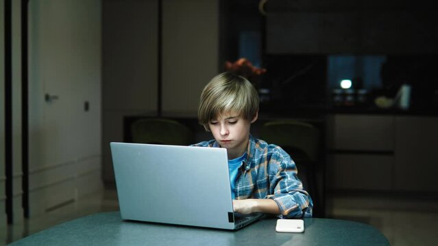 Serious concentrated focused school boy using laptop, doing homework, or playing computer games, sitting in the kitchen alone in the evening. Handsome caucasian teenager boy and laptopSerious concentr