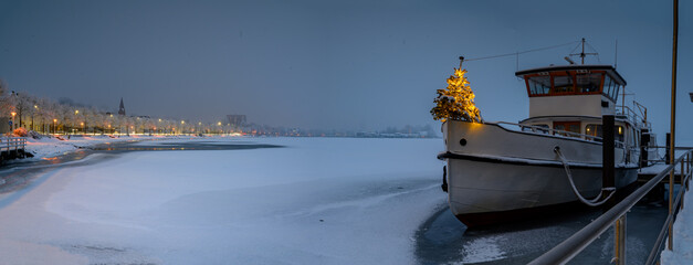 Ferry with Christmas tree standing at edge of bow. Panorama of ship boat moored at the pier covered with snow and promenade in winter in Schleswig. Winter, Christmas time on the Schlei, Schleswig. - obrazy, fototapety, plakaty