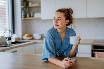 Young woman with smartphone enjoying cup of coffee at morning, in her kitchen.