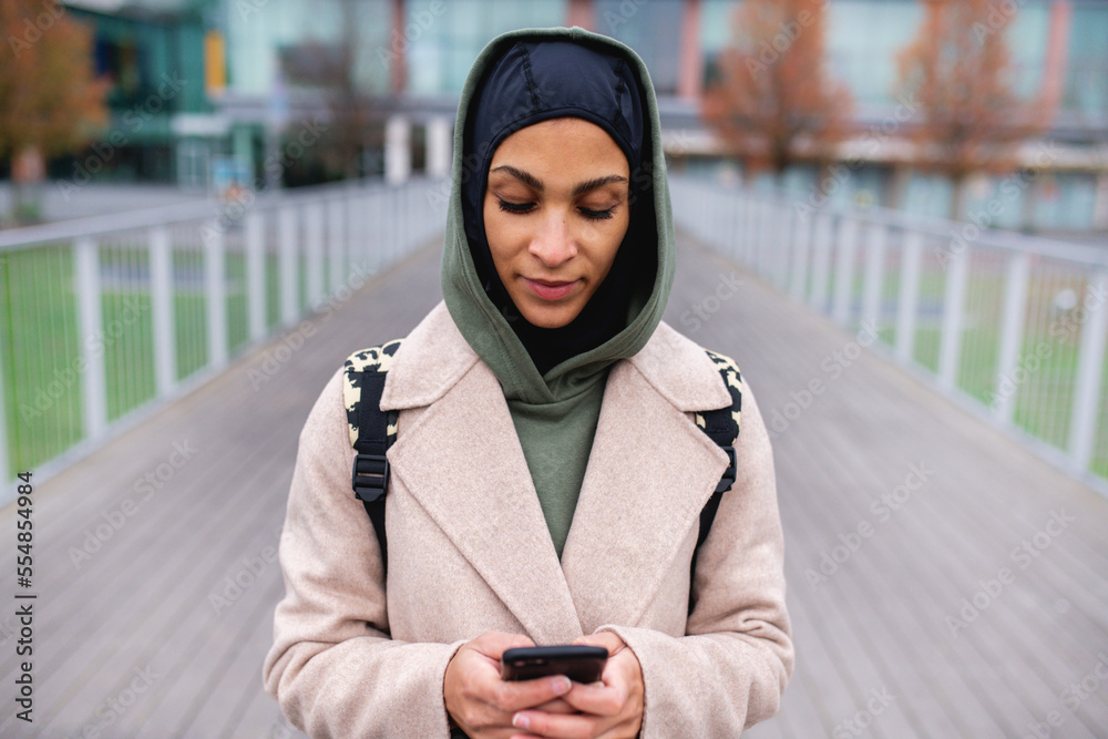 Wall mural portrait of young muslim woman in coat, standing outdoor in city, scrolling smartphone.