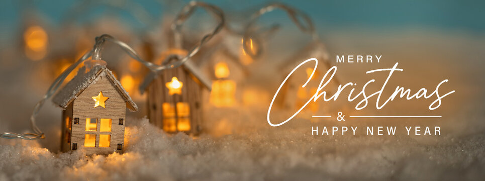 Christmas Greeting Card, English text. Abstract Christmas Winter Panorama with Wooden Houses Christmas String Lights in Cold Snow Landscape and Glowing Golden Lights in Background. Panorama, Banner. 