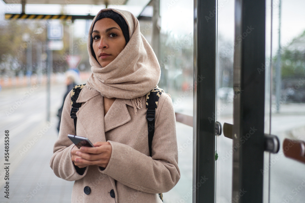 Wall mural young muslim woman with smartphone waiting for bus at city bus stop.