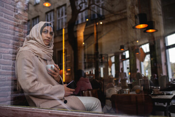 Young muslim woman with smartphone enjoying cup of coffe in cafe.
