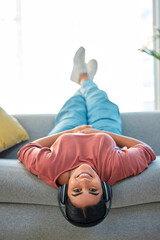 Woman, sofa and upside down with headphones, smile or music in portrait to relax in lounge. Happy lady, couch or audio for happiness, streaming podcast or internet radio in home living room in Mexico