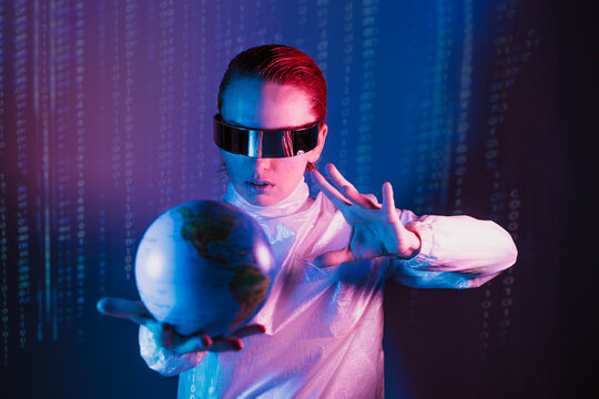 Concentrated woman in futuristic holographic clothes and VR glasses holding earth globe model on the one hand and affecting on it by another hand with matrix code background. Neon blue and pink light.