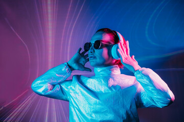 Portrait of concentrated woman in sunglasses and headphones in neon light. Music lover. Silent disco. Woman in holographic clothes on the background of music vibes background. Futuristic Party.