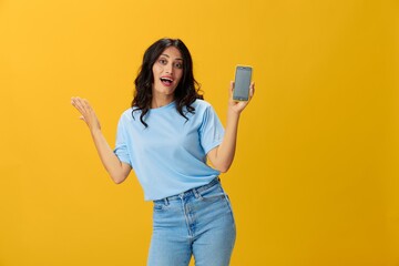 Woman blogger with a phone in her hands in a blue t-shirt and jeans on a yellow background smile signs gestures symbols, online communication and video call, copy space, free background