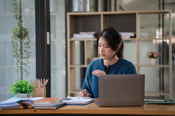 Fototapeta premium A beautiful Asian businesswoman analyzes charts using a laptop calculator at the office.