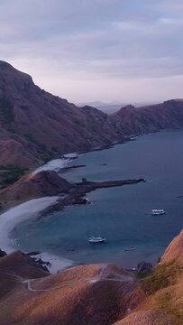 Vertical video of Aerial shot of steep rocky island at sunset with beach, boats in front