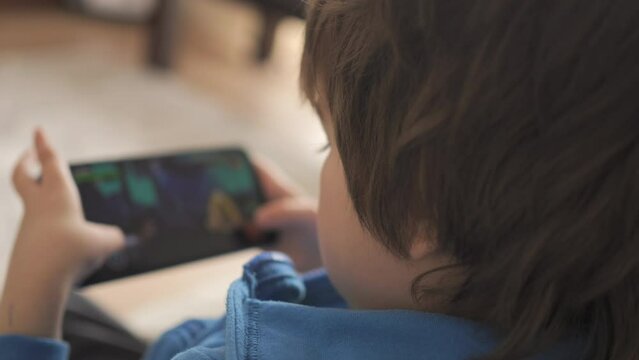Child Playing Games In Phone at Home Sitting on Couch. Boy Playing Video Game on Mobile Phone. Preschooler Plays Video Game Smartphone on Sofa. Kid Using Phone for Gaming Online Education Social Media