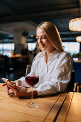 Vertical portrait of happy blonde young woman using smartphone, typing message sitting at table with glass of red wine at restaurant with dark luxury interior. Happy lady chatting with boyfriend.