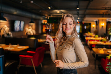 Portrait of elegant blonde woman holding glass of red wine standing in restaurant with luxury...