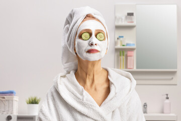 Woman in a bathrobe with a face mask with cucumber on eyes standing in a bathroom
