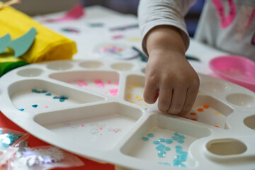 Fototapeta na wymiar a child's hand sorts shiny colored pieces for decoration in a white paint palette