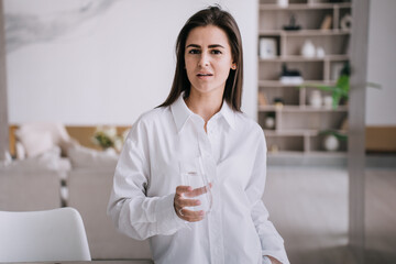 Attractuve Italian brunette young adult woman in white shirt holds glass of water looks at camera with confident face standing against blurry living room. Healthy lifestyle and women domestic leisure.