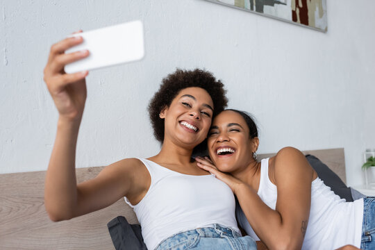 happy african american lesbian couple taking selfie on smartphone in bedroom