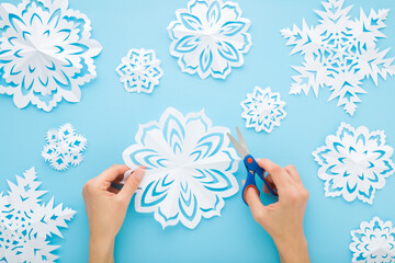 Young adult woman hands cutting different white snowflake shapes from paper on light blue table...