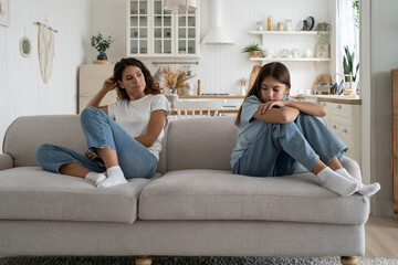 Upset teenage daughter and young woman mother sitting separately in silence on sofa at home,...