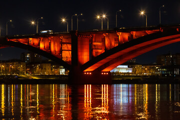 Fototapeta premium Night landscape with red illuminated of Communal Bridge in Krasnoyarsk, Russia. View of the Yenisey River at night. Reflection of lighting lights in the rippled water surface