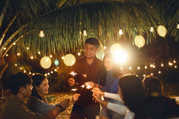 Happy Asian couple with firework sparkler in party anniversary with group friends in celebrating under light at night. Friendship and celebration concept.