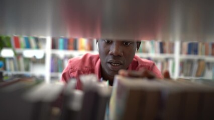 Positive fussy African man choosing book in library standing near rack and moving fingers along shelf. Optimistic inquisitive black guy student in headphones selects textbooks to prepare for exams