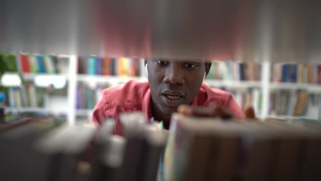 Positive fussy African man choosing book in library standing near rack and moving fingers along shelf. Optimistic inquisitive black guy student in headphones selects textbooks to prepare for exams