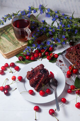 Chocolate cake with sweet cherry, cup of tea on vintage book and branch of blue flowers on white background..