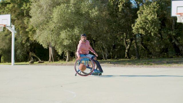 Strong man in sports wheelchair playing basketball at open-air court on sunny day.Hispanic man in backward cap and hoodie riding on circle, dribbling ball and throwing it in hoop.Amputee sport concept