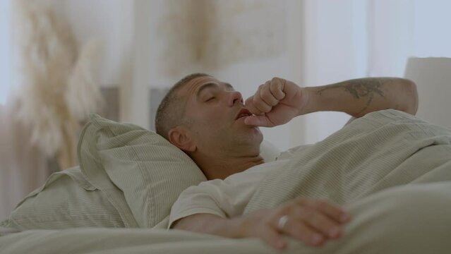 Peaceful man lying in bed, waking up, stretching and yawning. Medium shot of handsome man waking up in early morning, preparing for getting out of bed, relaxing at home. Sleep cycle, health concept