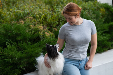 Caucasian red-haired woman cuddling with pappilion dog outdoors. Black and white continental spaniel.