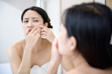 Obraz na płótnie Canvas Problem skin. Concerned young asian women popping pimple on cheek while standing near mirror in bathroom. young asian women with acne