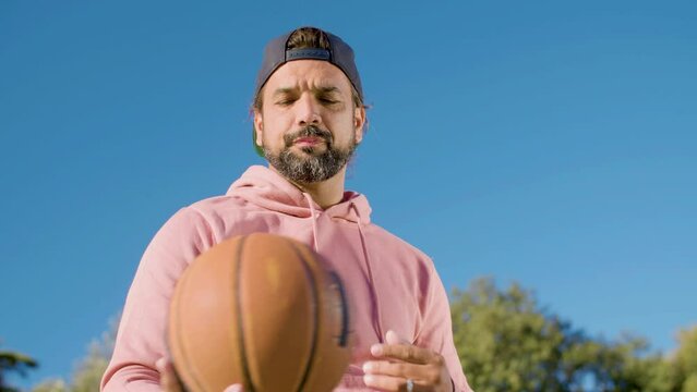 Bottom shot of basketball player throwing ball outside against blue sky background. Bearded Hispanic man in backward cap and pink hoodie playing ball game on sunny day. Sport, active lifestyle concept