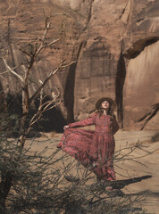 A girl wearing a long red dress and a straw hat stands in the windy and rocky AlUla, desert, Saudi Arabia. 