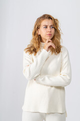 A young blonde girl with curly hair in a white knitted suit stands thoughtfully on a white background