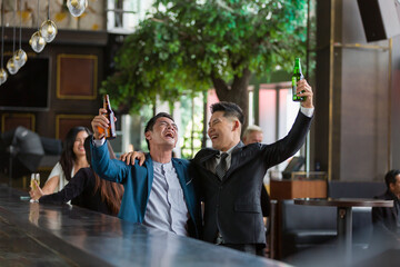Two Asian business man or partnership holding up bottles of beer and drinking discussing project work in the office work space