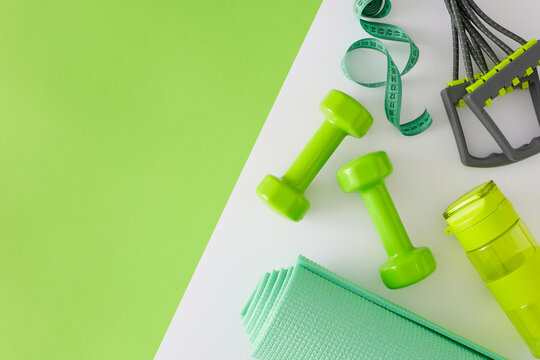 Fitness concept. Flat lay photo of dumbbells exercise mat bottle of water elastic expander on white and green background with copy space. Minimal sports accessories idea.