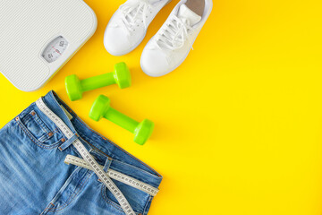 Slimming concept. Top view photo of blue jeans with tape measure, scales, dumbbells and white sports shoes on yellow background. Flat lay with copy space.