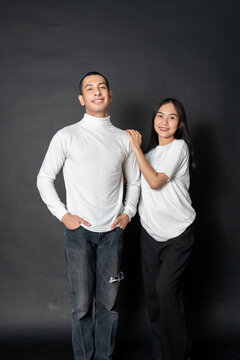 Young Energetic Couple Wearing White Blank Shirt Doing A Pose While Smiling Happily With A Black Background