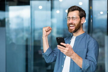 Obraz na płótnie Canvas Businessman in shirt near window using phone , mature man with beard received notification online good news, worker celebrating victory rejoices, holding hand up gesture of triumph and achievement.