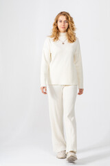 A young blonde girl with curly hair in a white knitted suit smiles and steps on a white background