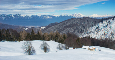 Fototapeta na wymiar Postcard panoramic landscape. Small stone houses amid snowdrifts with mountains in the background. Photographed in Combai, Treviso, Italy.