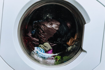 Wet dirty clothes are washed in a washing machine inside with water behind a glass round porthole. Photography, laundry.