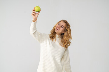 A young pretty curly blonde woman holds a yellow apple in her hands. The girl looks happy and smiles, she is dressed in a white knitted suit. Near the model there is a lot of space for a