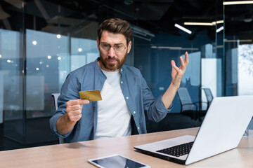 Cheated and disappointed businessman working inside office, man holding bank credit card and trying...