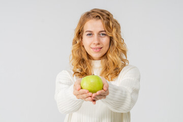 A young pretty curly blonde woman holds a yellow apple in her hands. The girl looks happy and smiles, she is dressed in a white knitted suit. Near the model there is a lot of space for a