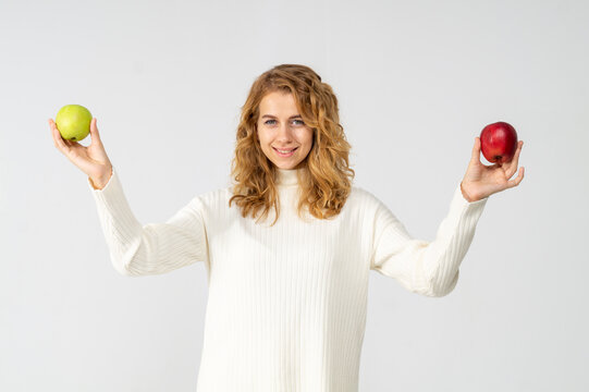A young pretty curly blonde woman holds a yellow green and red apple in her hands. The girl looks happy and smiles, she is dressed in a white knitted suit. Near the model there is a lot of space for a