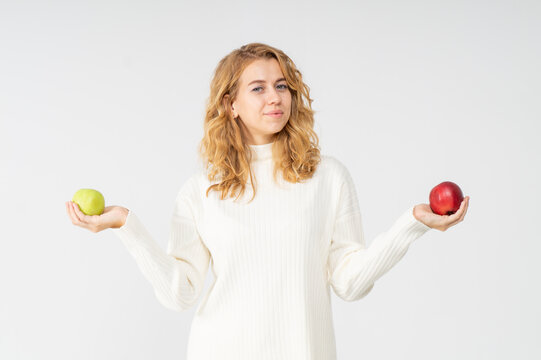 A young pretty curly blonde woman holds a yellow green and red apple in her hands. The girl looks happy and smiles, she is dressed in a white knitted suit. Near the model there is a lot of space for a